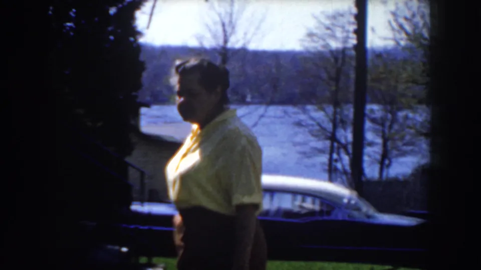 Closer 1959 Michigan home-movie frame showing the woman in profile near a dark sedan, with trees, steps, railing, and water behind her.