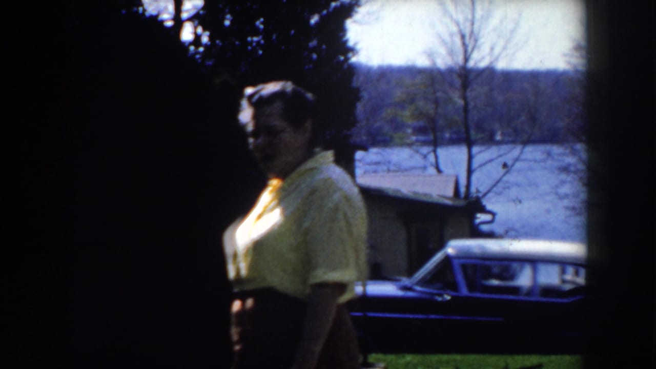 Woman in a yellow shirt walks in a shaded yard with a dark car and water behind her.