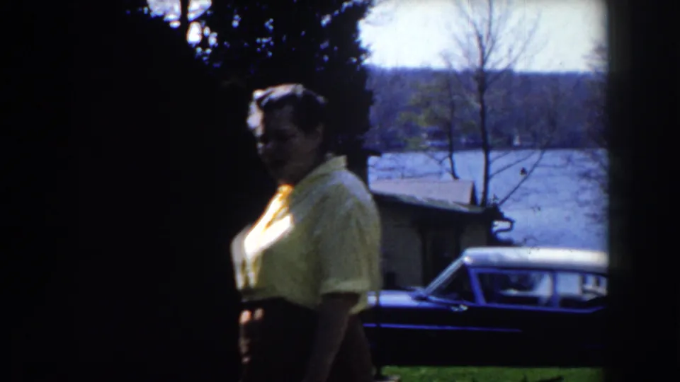 In Michigan 1959 home-movie footage, a woman in a yellow shirt walks past a dark car with trees and water in the background.