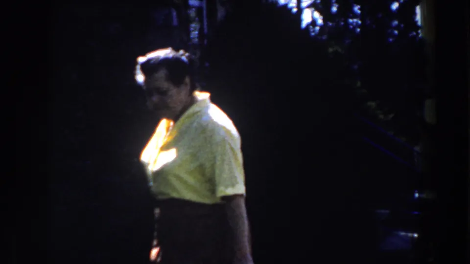 A woman walks through a shaded Michigan yard, her yellow shirt catching strong sunlight in the 1959 8mm footage.