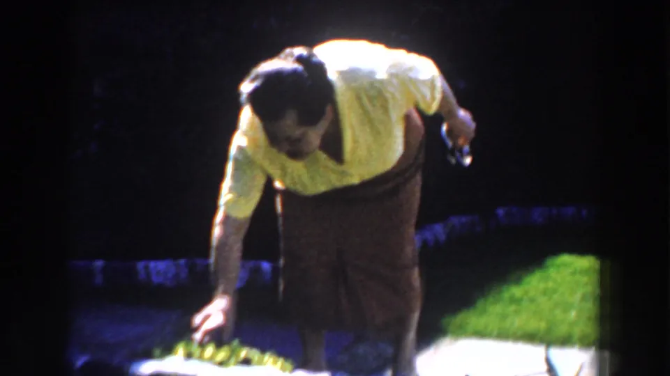 The woman bends toward a low stone border in a sunlit yard, with grass and pavement visible in the 1959 home movie frame.