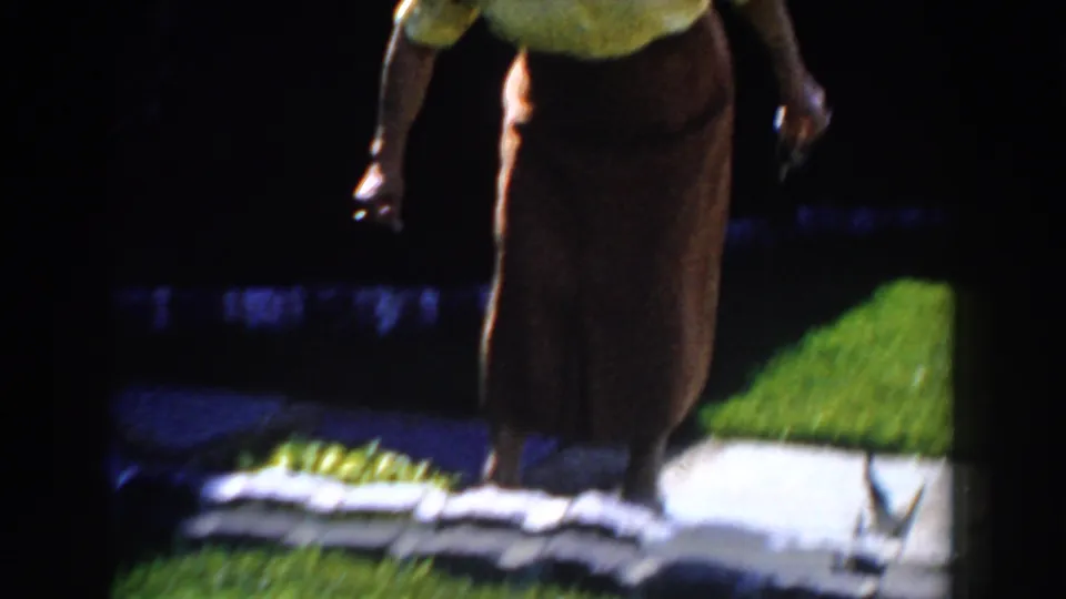 A woman stands beside a low stone garden border in a Michigan yard, with bright grass and walkway visible in the 1959 8mm frame.