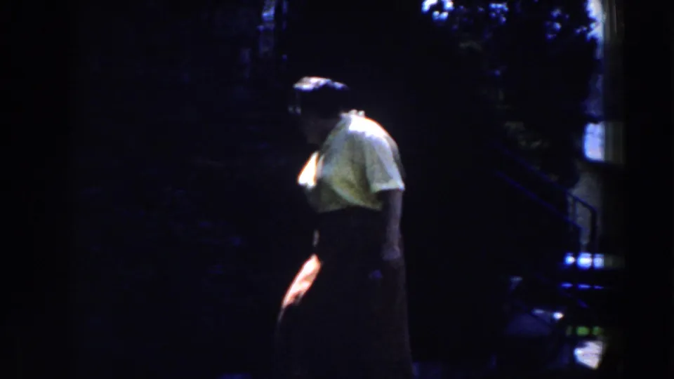 In 1959 Michigan home-movie footage, an adult woman in a yellow shirt and dark skirt stands beside dark hedges.