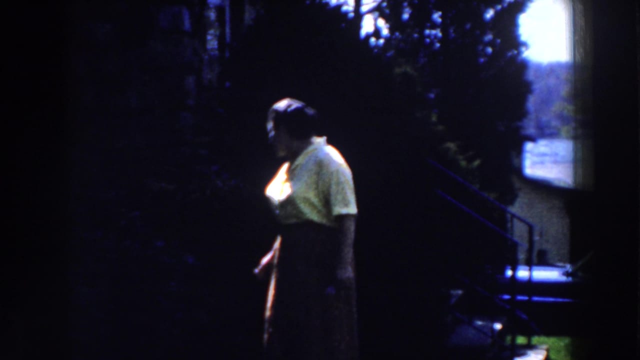 Woman in yellow shirt stands in profile beside dark foliage.