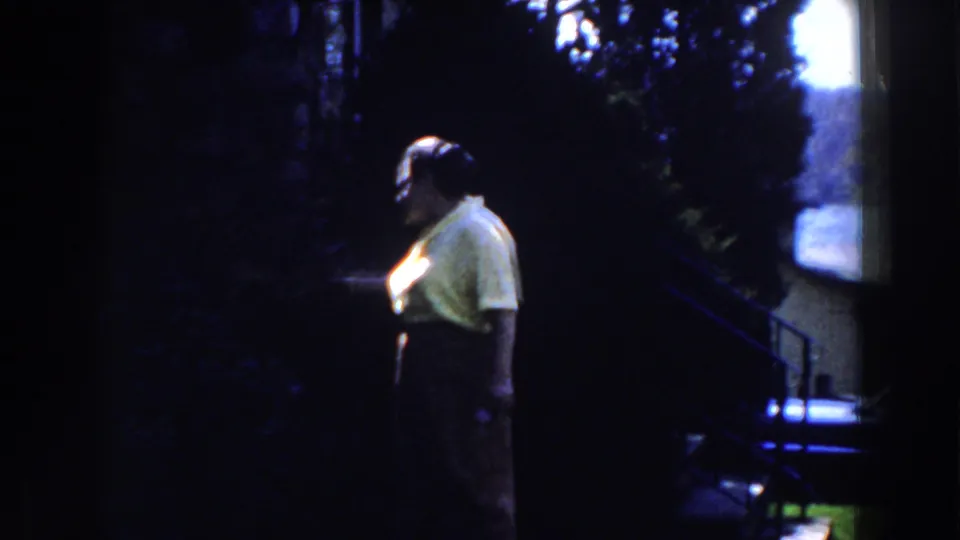 The woman in yellow turns slightly in the dim outdoor frame, with hedges and a bright background opening behind her.