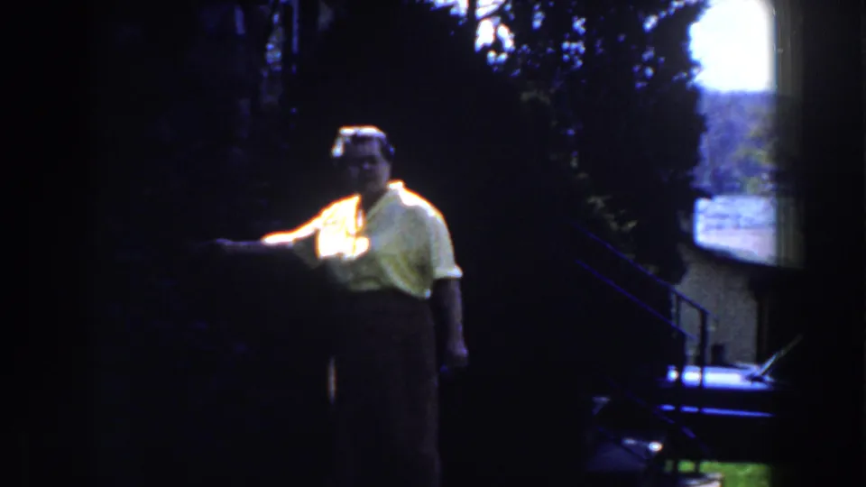 1959 Michigan 8mm frame of a woman in a yellow blouse and dark skirt pointing in a backlit wooded outdoor setting.