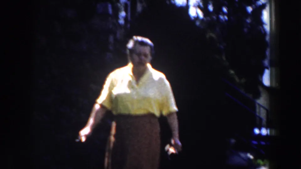 1959 Michigan home-movie frame showing a woman in a yellow blouse and dark skirt standing in strong backlight near trees.