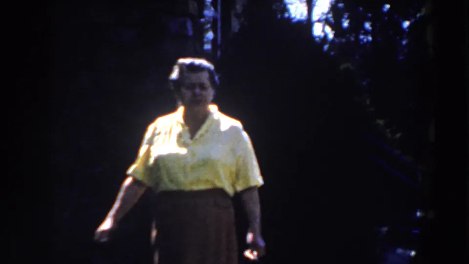 1959 Michigan 8mm frame of a woman in a yellow blouse moving forward in a shaded wooded area with a stair rail nearby.