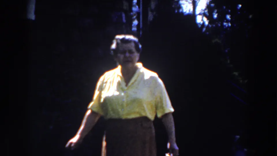 1959 Michigan home-movie frame showing a woman facing the camera in a yellow blouse, with trees and deep shade behind her.