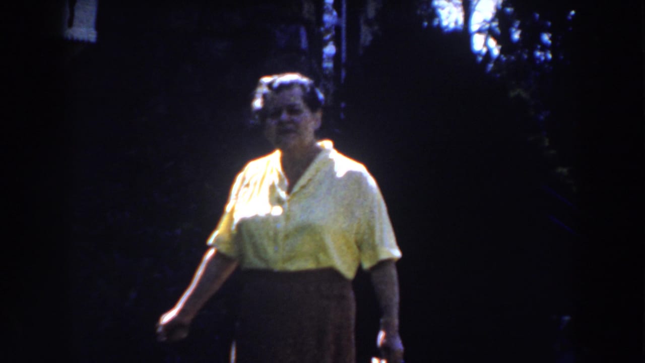 In 1959 Michigan home-movie footage, an adult woman in a yellow blouse and dark skirt walks through a dim outdoor setting.