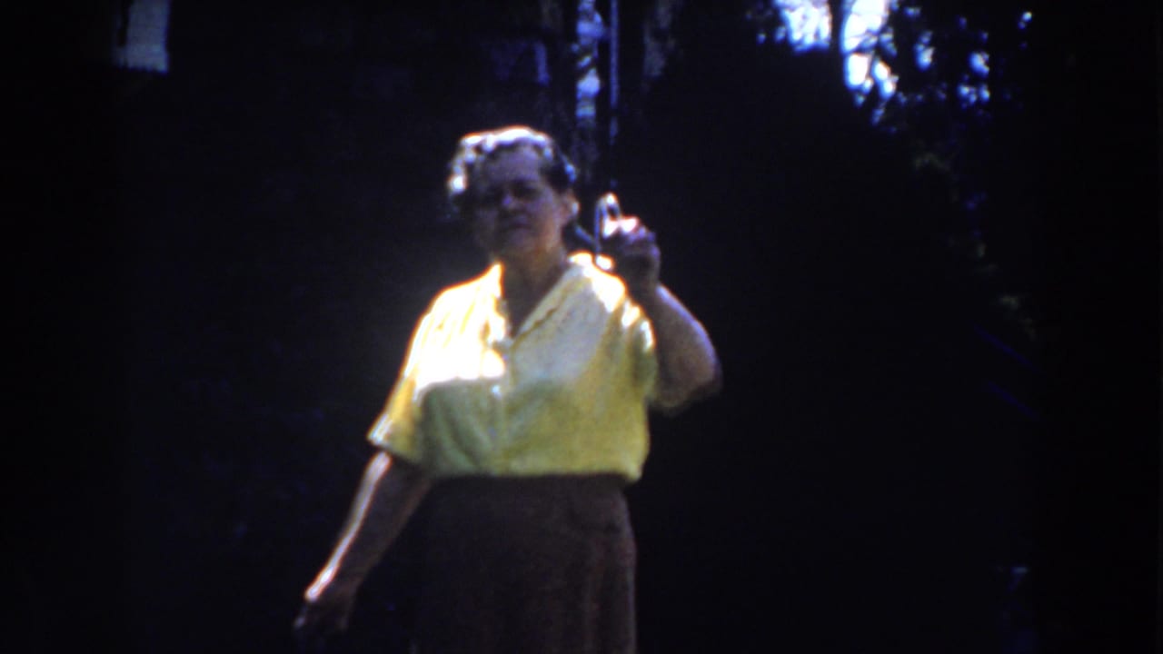 Adult woman in a yellow blouse raises one hand while facing the camera outdoors.