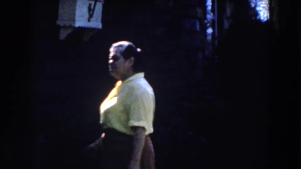 The 1959 Michigan home movie shows the woman in profile, walking past a dark wall or shaded exterior space.