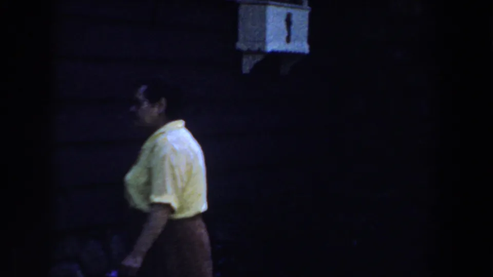 The woman continues across the frame in 1959 Michigan footage, partly silhouetted by deep shade around a dark exterior wall.