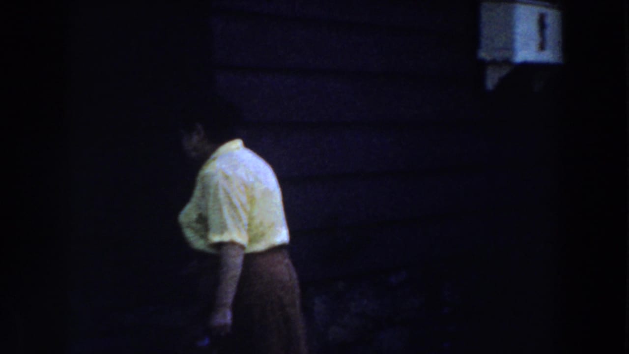 In a 1959 Michigan home movie, a woman in a yellow blouse and dark skirt moves toward a shadowed house entrance at night.