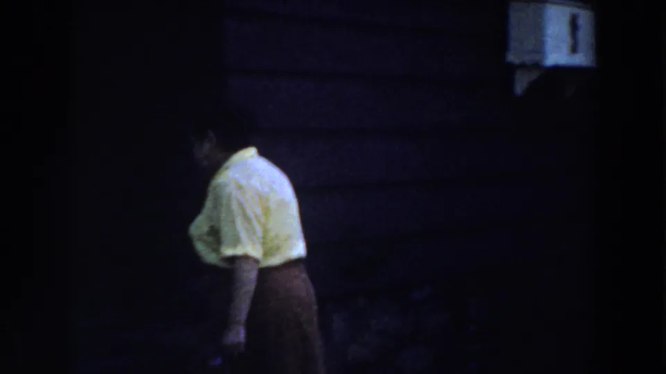 In a 1959 Michigan home movie, a woman in a yellow blouse and dark skirt moves toward a shadowed house entrance at night.