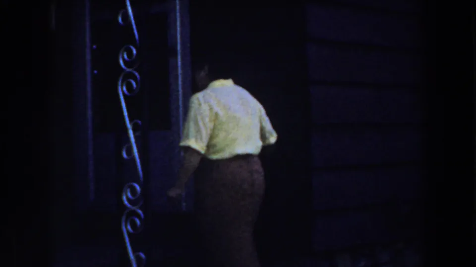 The woman approaches a dark porch doorway with curved metalwork visible along the entrance area.