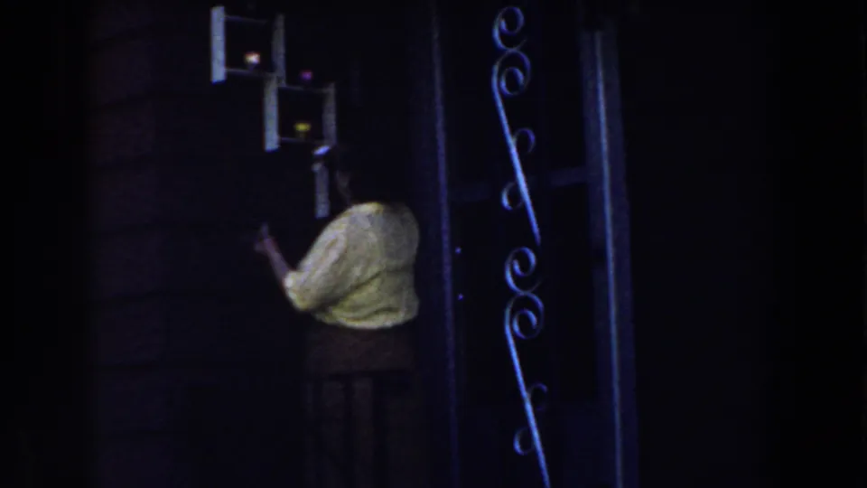 Near the end of the 1959 Michigan clip, the woman stands by a dark doorway beneath small porch fixtures and decorative metalwork.