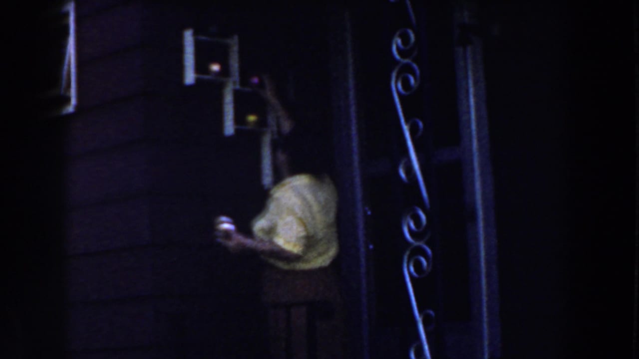 Woman in a light blouse reaches toward a small wall cupboard in a dim interior.