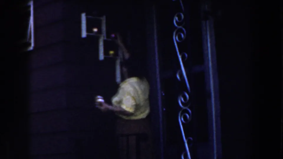 1959 Michigan 8mm frame showing a woman from behind reaching up toward a small wall cupboard in a dark home interior.