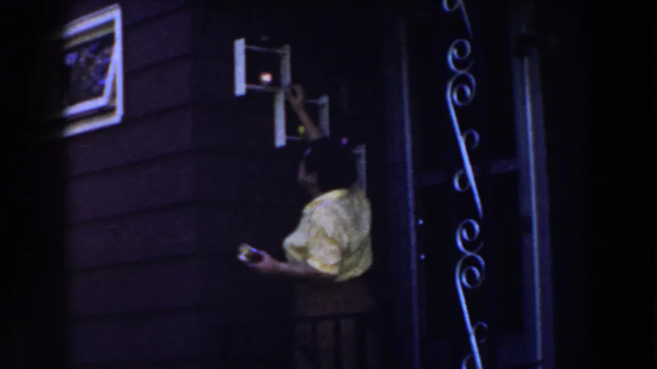 1959 Michigan home-movie frame showing a woman beside a wall cupboard, framed picture, and decorative wrought-iron detail.
