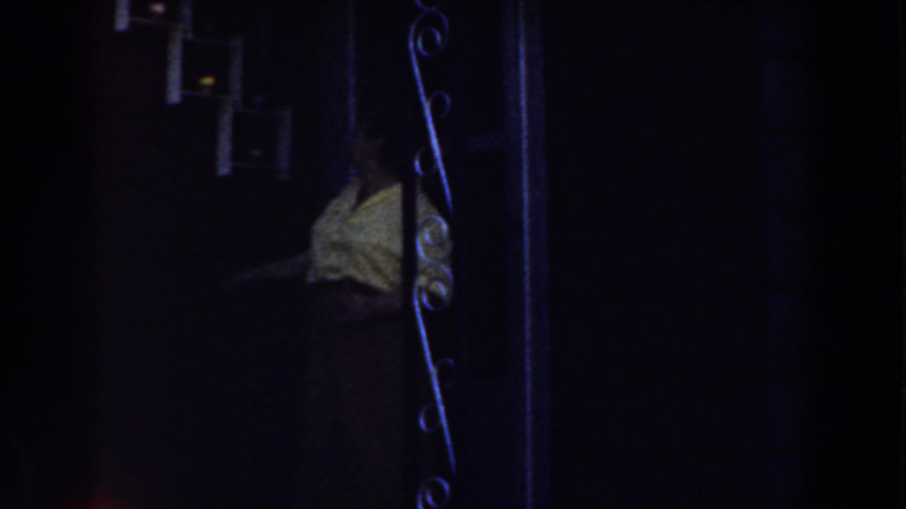 Woman in a yellow shirt partly obscured by a dark doorway and wrought iron gate.