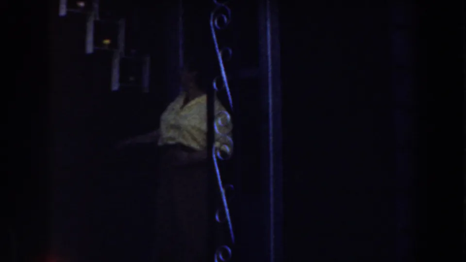 In 1959 Michigan home-movie footage, a woman in a yellow shirt stands near a dark doorway behind wrought iron scrollwork.