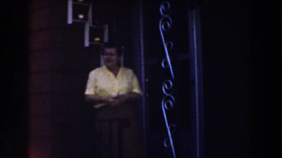 A woman in a yellow blouse faces toward the camera in dim 1959 Michigan home-movie footage near a brick doorway.