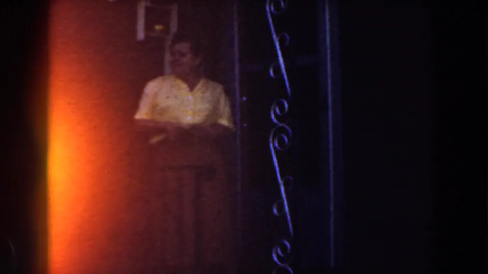 Adult woman in a yellow shirt stands by a doorway in 1959 Michigan home-movie footage, partly obscured by dark frame edges and warm exposure.