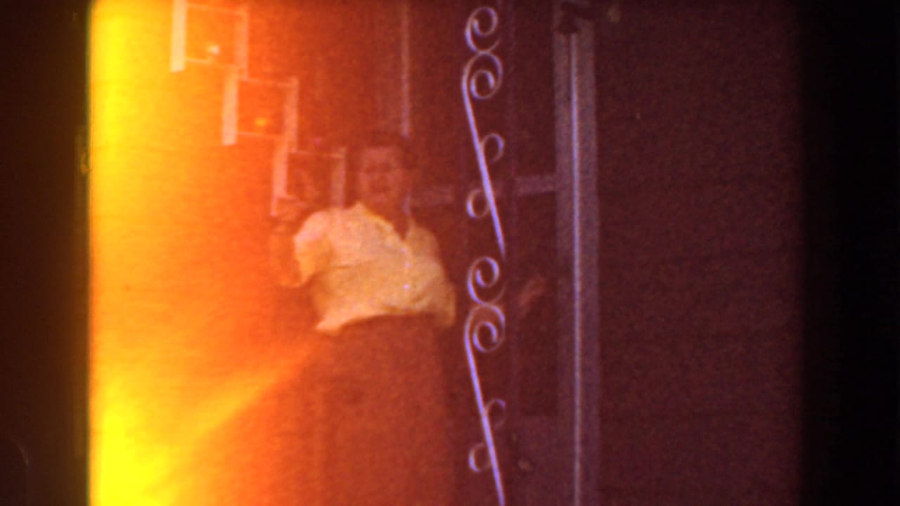 Woman in a yellow shirt waving beside a doorway and orange-lit wall.