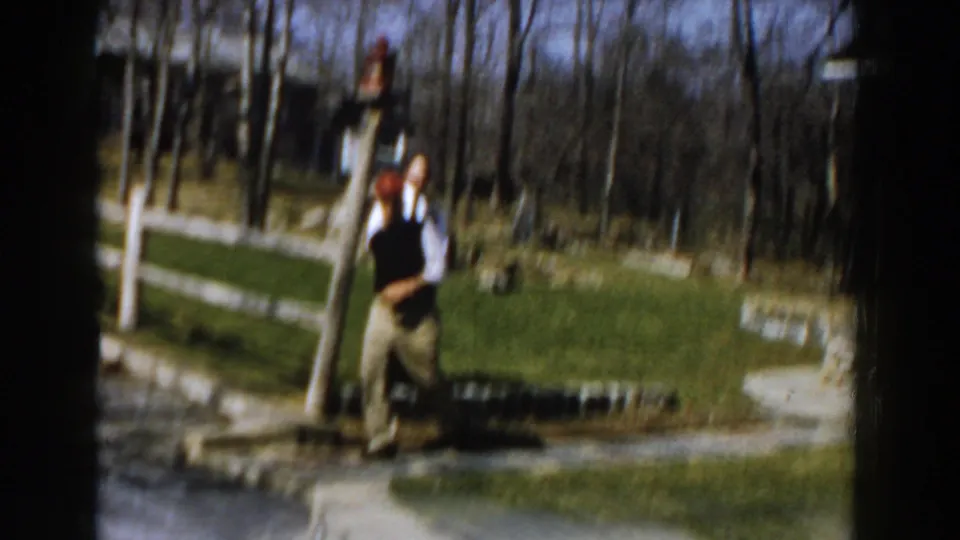 A man in a red cap, white shirt, and dark vest stands in a suburban yard, with bare trees and lawn behind him.