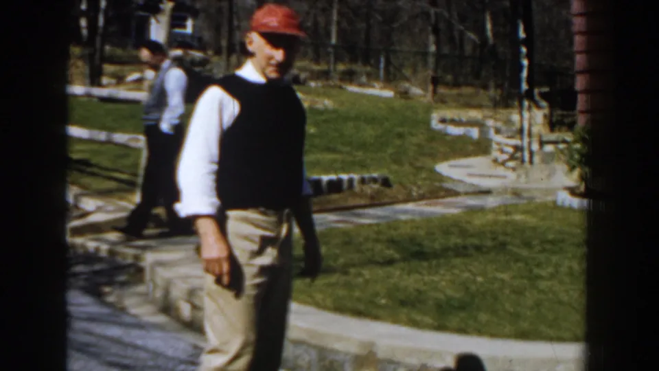 A man in a red cap and dark vest walks through a Michigan yard, with another adult figure visible behind him near paths and lawn.