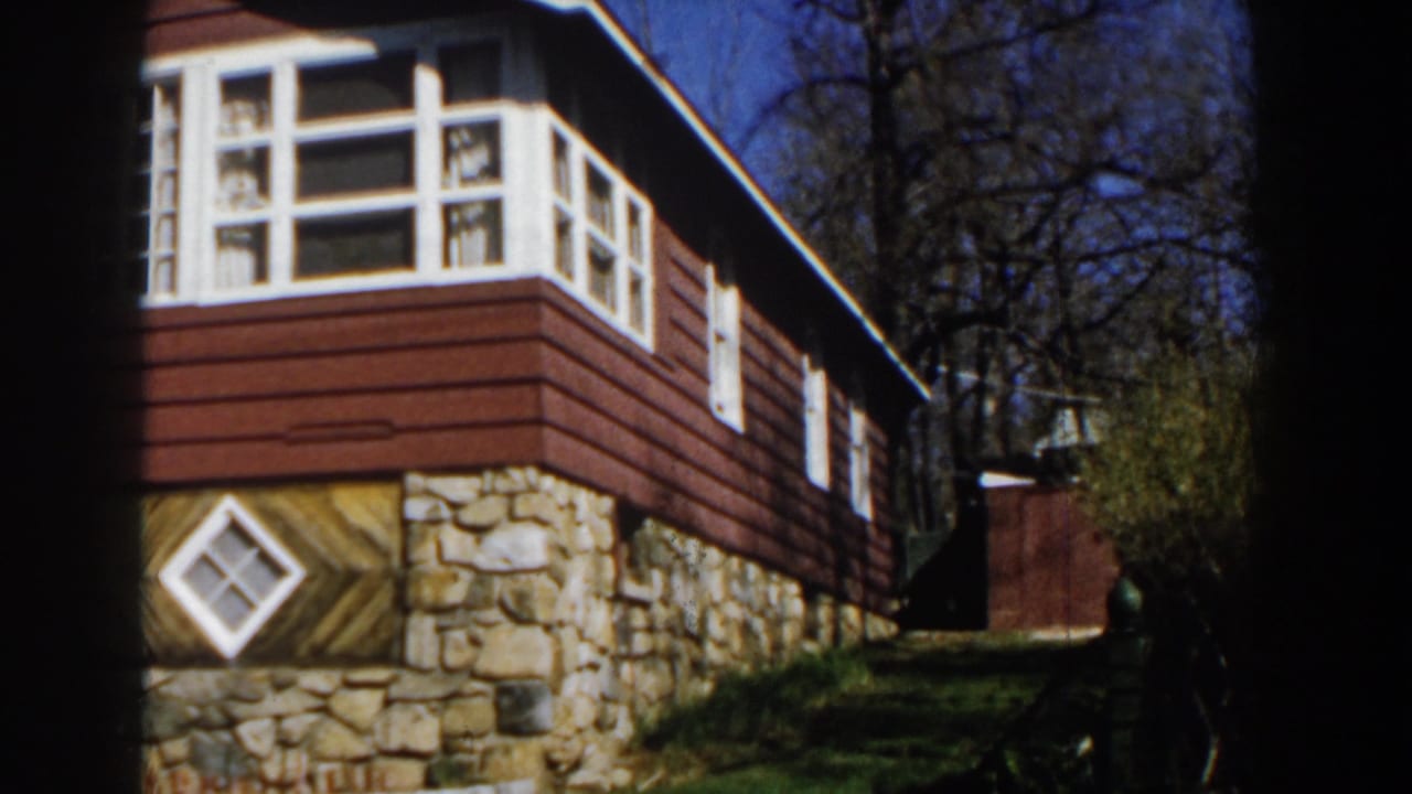 1959 Michigan 8mm frame showing a red cabin exterior with white trim, stone base, diamond window, bare trees, and blue sky.