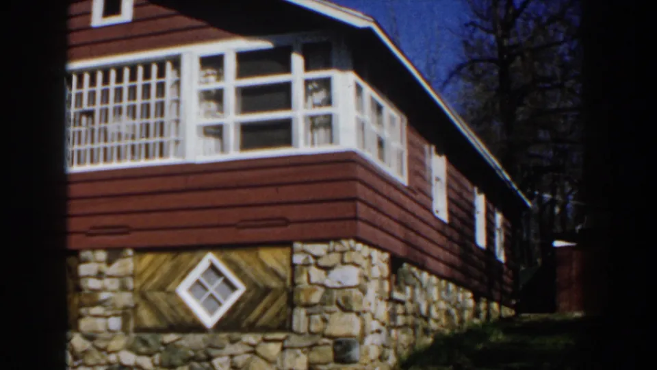 1959 Michigan home-movie frame centered on the cabin's white gridded upper window, red siding, stone base, and lower diamond window.