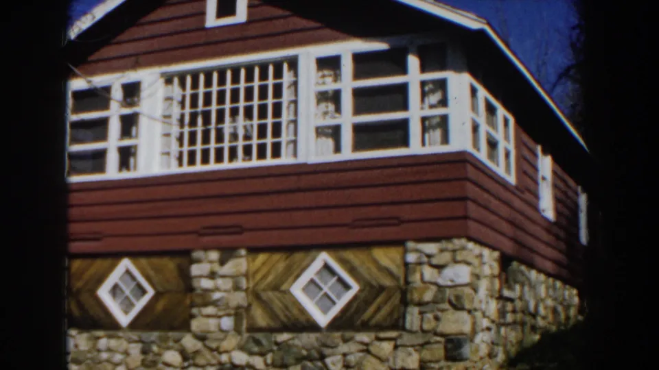 1959 Michigan 8mm frame with a closer view of the cabin facade, including red siding, white trim, gridded upper windows, and two lower diamond windows.