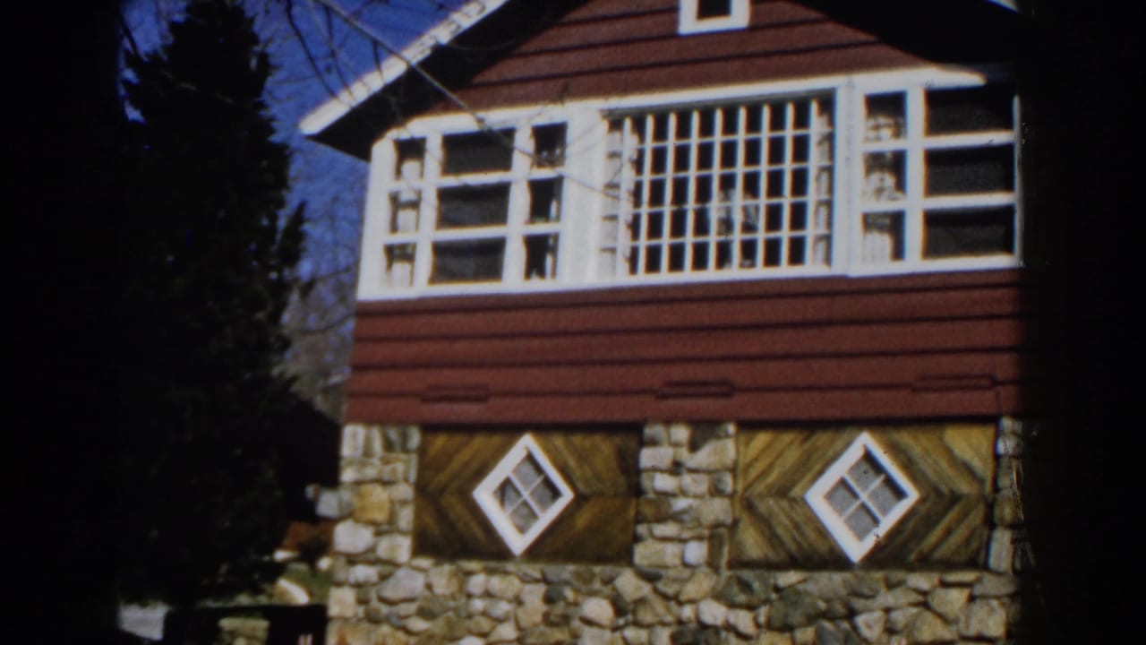 House corner with red siding, white windows, stone base, and trees behind it.