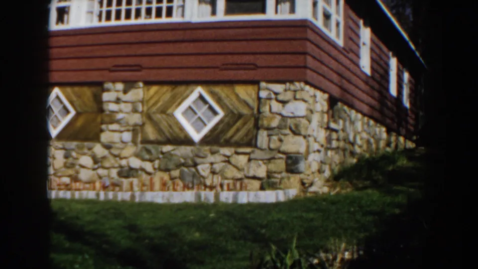 1959 Michigan home-movie frame showing the side of the red and stone house with grass in front.