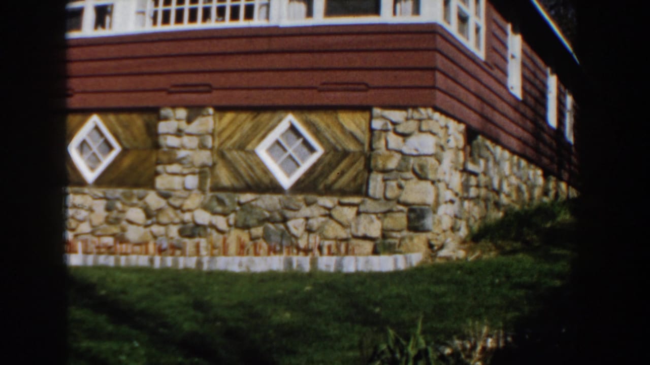 1959 Michigan 8mm frame showing a house exterior with red siding, stone lower facade, diamond windows, and grass lawn.