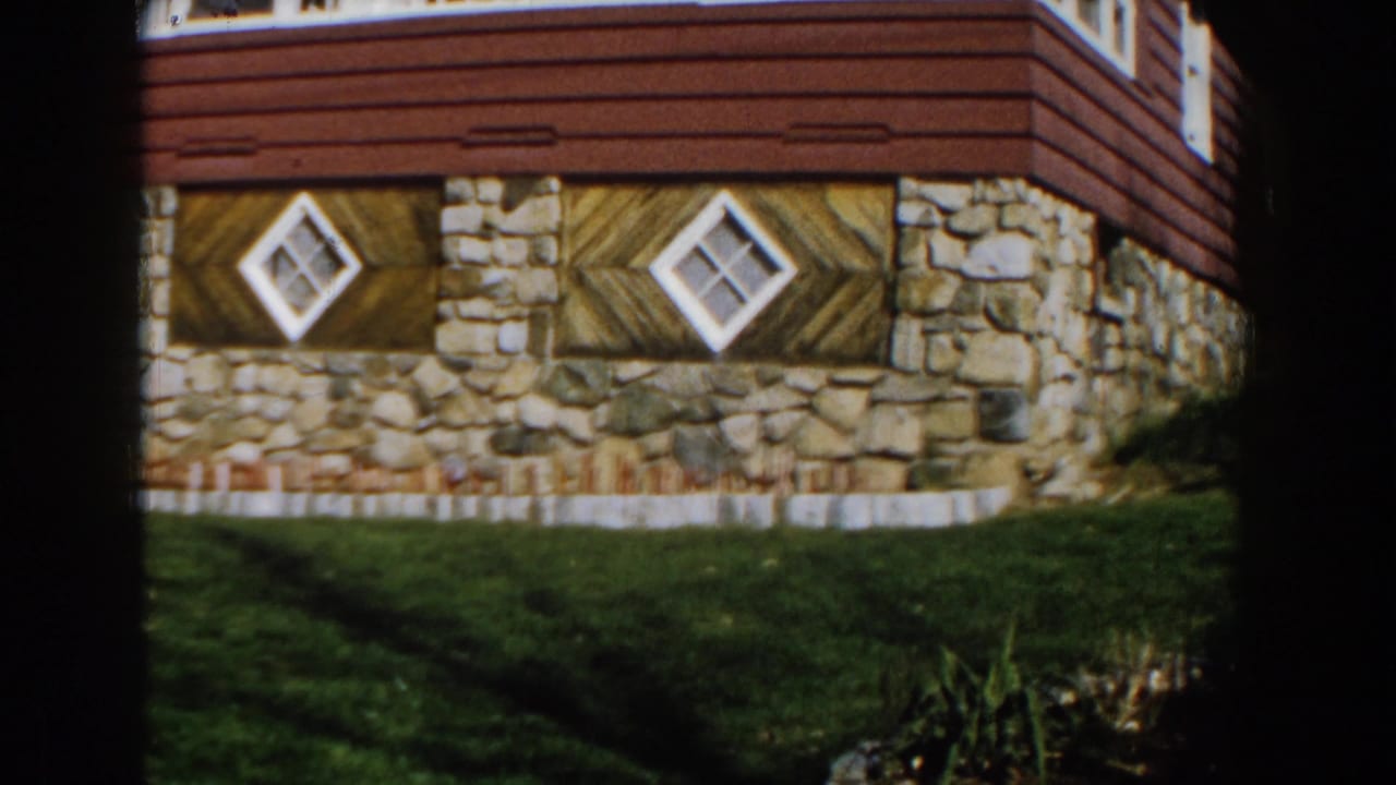 1959 Michigan home-movie view of a stone and red-sided house facade with two diamond windows above a lawn.