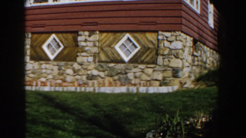 1959 Michigan home-movie view of a stone and red-sided house facade with two diamond windows above a lawn.