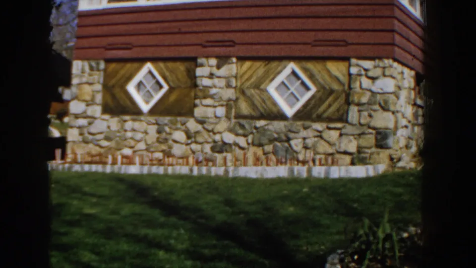 1959 Michigan exterior frame showing the lower wall of a house with stonework, red siding, diamond windows, and lawn shade.