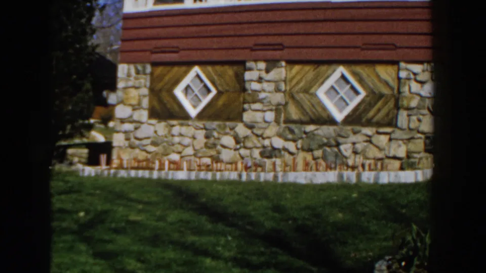 1959 Michigan 8mm frame of a red-sided house with a stone lower facade, patterned wood panels, diamond windows, and lawn.