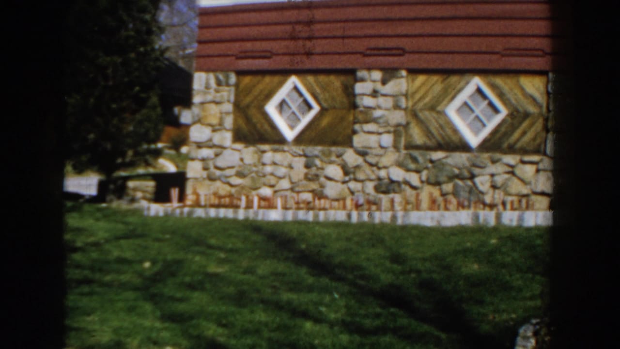 Stone-and-wood house facade with diamond windows, red siding, green lawn, and tree shadows.