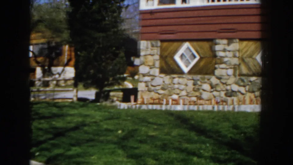 1959 Michigan home-movie frame with the house exterior shifted right, showing stonework, a diamond window, red siding, lawn, and nearby trees.