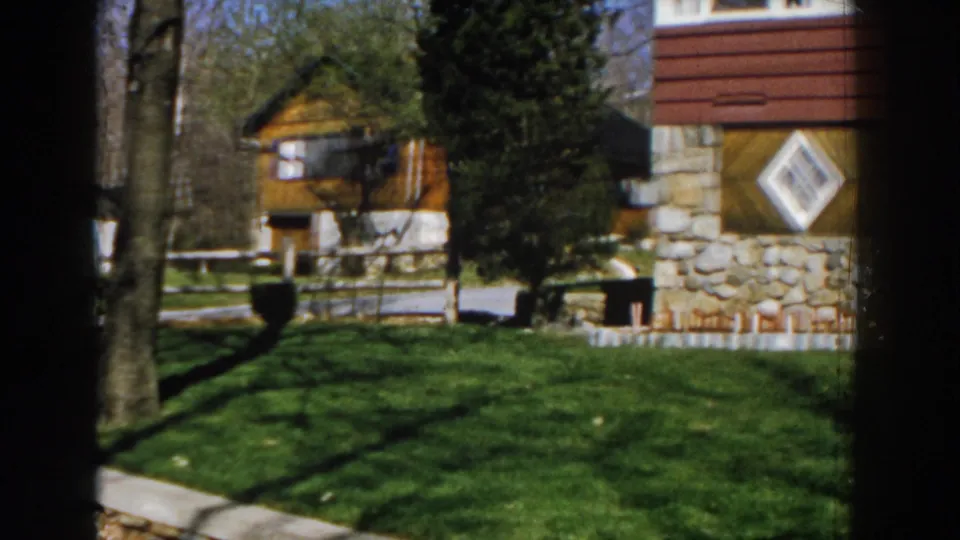 1959 Michigan 8mm frame showing a small wooden building beyond the lawn, tree trunks, white fencing, and the stone house edge.