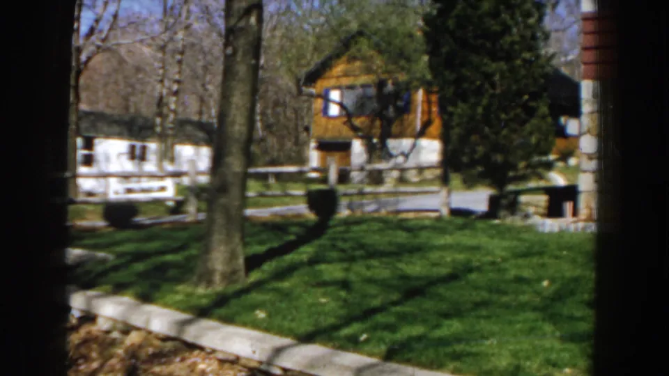 1959 Michigan frame looking across a green lawn toward a wooden building and white fence, with tree trunks, an evergreen, and stone edging in view.