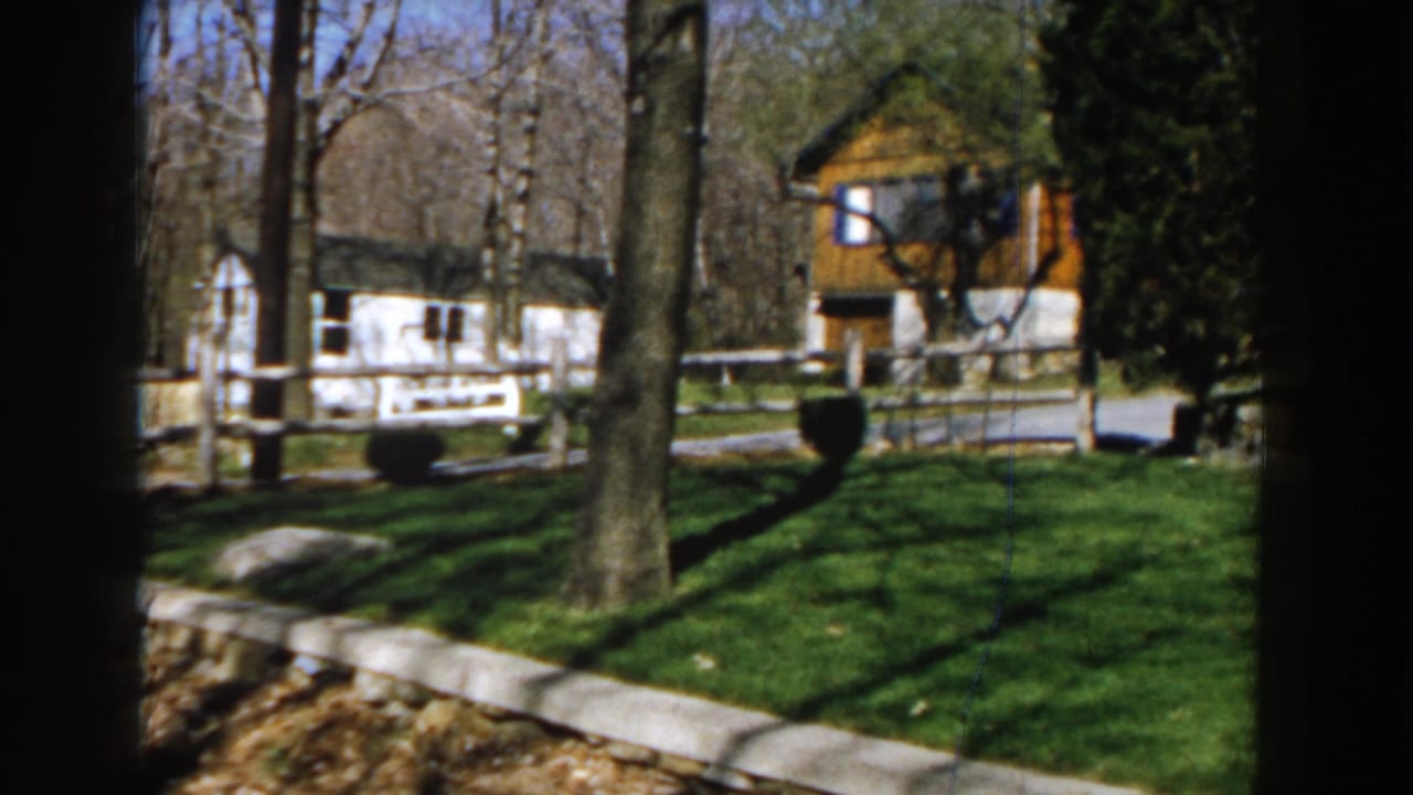 Rural yard with split-rail fence, trees, white building, and rustic wood structure in the background.