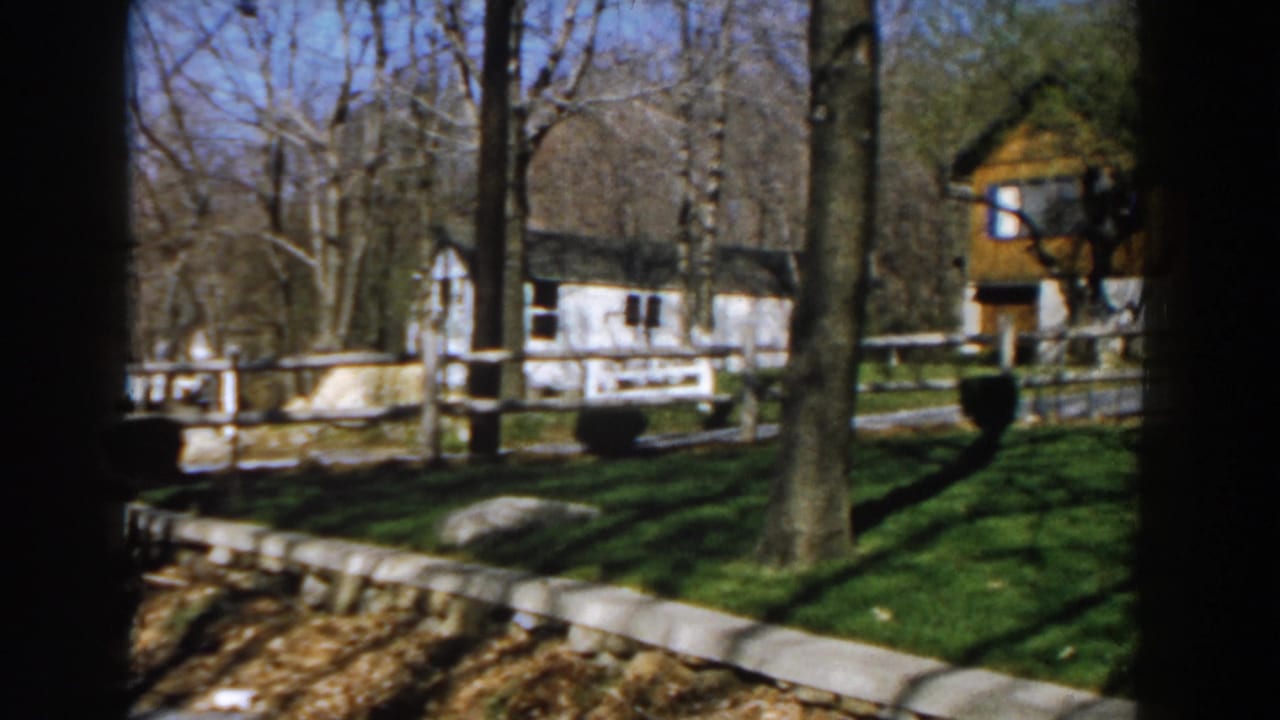 Split-rail fence and grassy yard with a white building and wood-sided structure beyond trees.