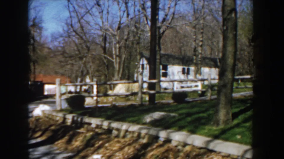 1959 Michigan home-movie view of a white rural building behind fencing, with bare trees, lawn, stone edging, and a narrow channel in front.