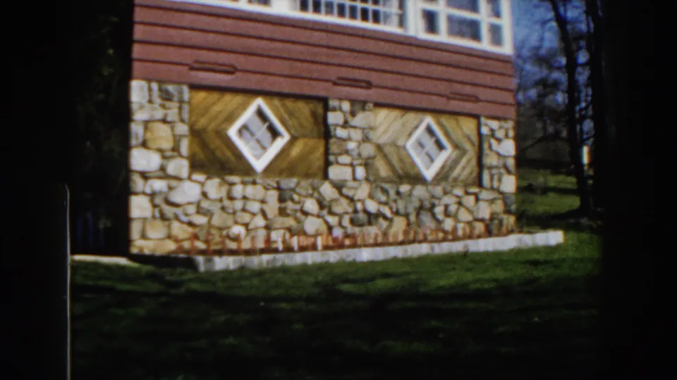 1959 Michigan frame showing a rustic building exterior with stone foundation walls, diagonal wood siding, diamond windows, grass, and trees.