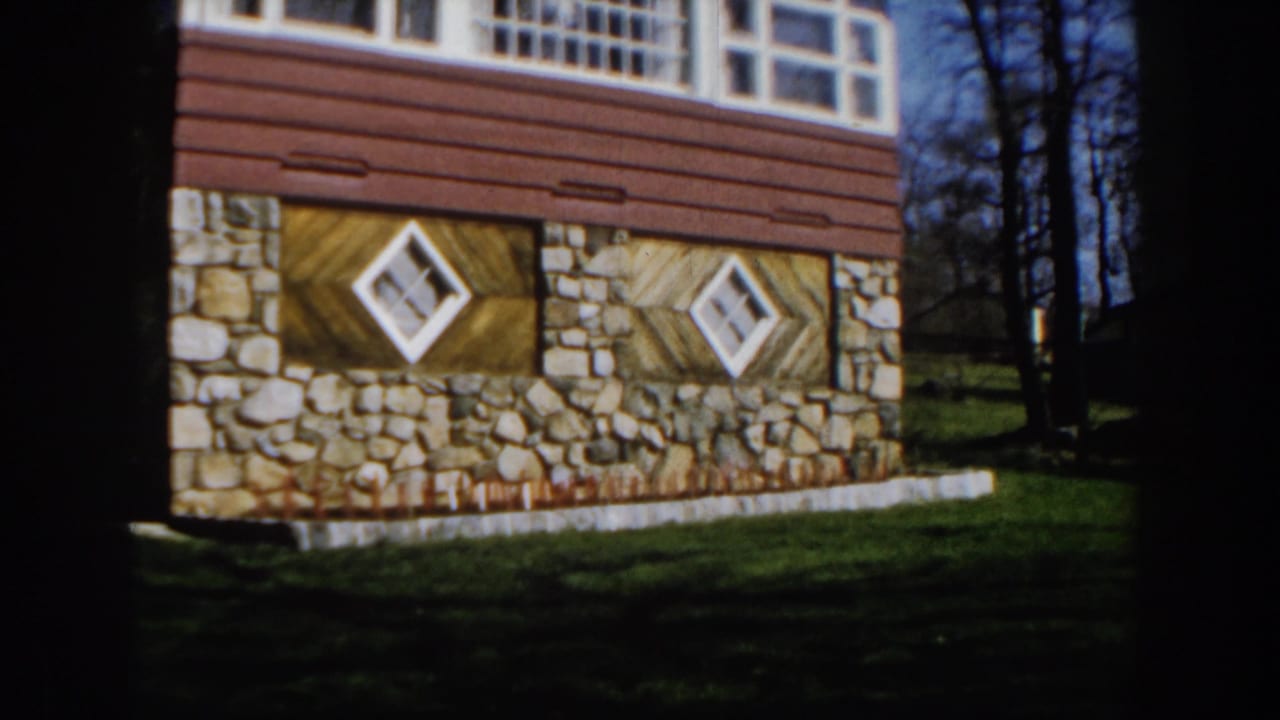 1959 Michigan home-movie frame showing a house with stone foundation, wood siding, diamond windows, grass, and bare trees.